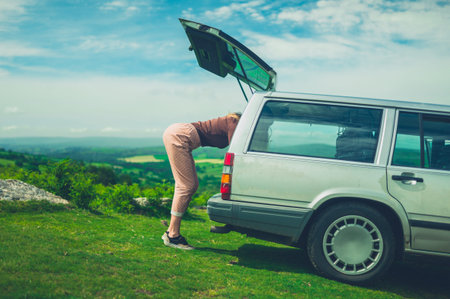 A young woman is looking for something in the trunk of her car in nature on a sunny summer day