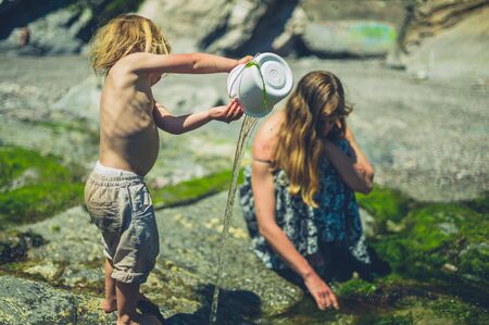 A little toddler is on the beach with his mother and is splashing water from a bucketの写真素材