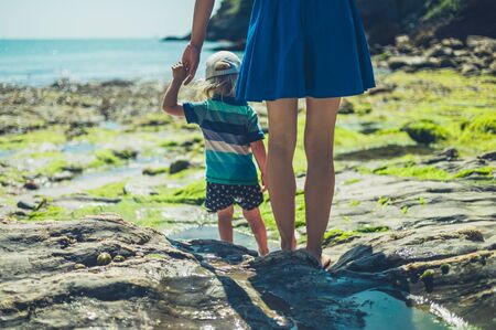 A young mother is walking on a rocky beach with her toddler on a sunny summer dayの写真素材