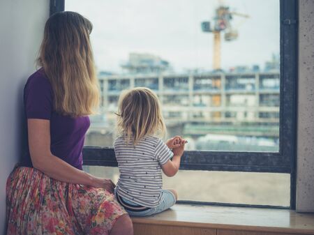 A young mother and her little toddler are looking out the window of a city apartmentの写真素材