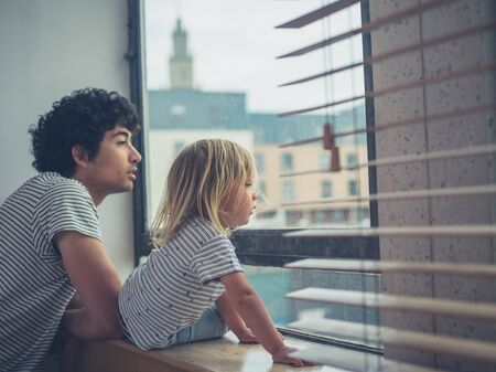 A young father and his toddler are looking out the window of a city apartmentの写真素材