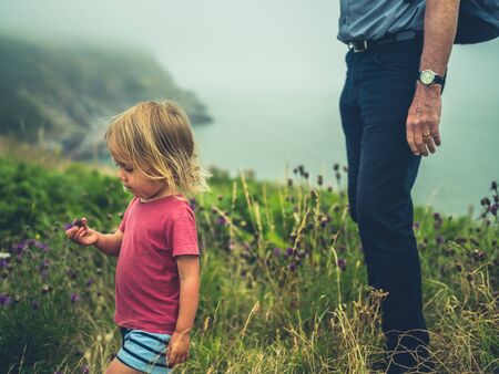 A little toddler is walking by the coast with his grandfather on a cloudy day. Authentic family lifestyle shot.の写真素材