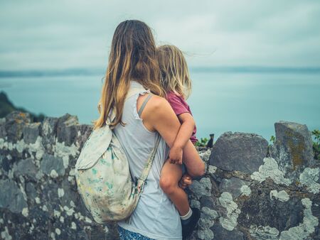 A young mother and her toddler are standing by a wall on the coastの写真素材