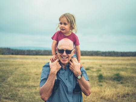 A little toddler is riding on the shoulders of his grandfather outdoors in nature on a summer dayの写真素材
