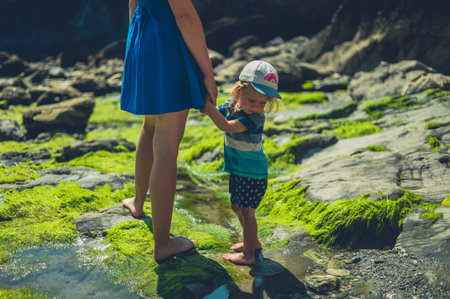 A young mother is walking on a rocky beach with her toddler on a sunny summer dayの写真素材