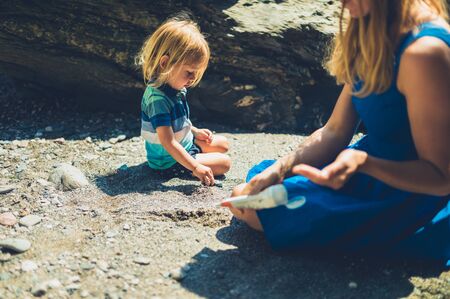 A young mother is on the beach with her toddler and is applying suncream to his skinの写真素材
