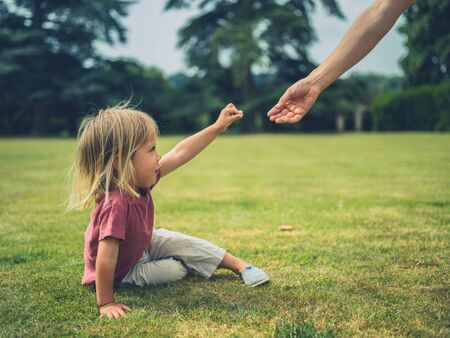 A little toddler is sitting on the grass in a park and is offering his mother a flowerの写真素材
