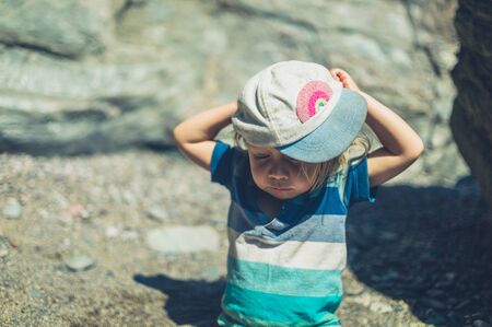 A little toddler is sitting on the beach and is putting on his hatの写真素材