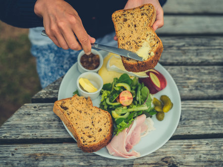 A young woman is buttering bread outdoors at a picnic table to make a sandwichの写真素材