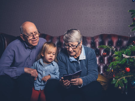 A little toddler and his grandparents are sitting on a sofa looking at a smartphone by the christmas treeの写真素材