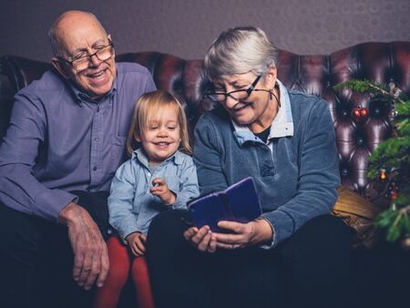 A little toddler and his grandparents are sitting on a sofa looking at a smartphone by the christmas treeの写真素材