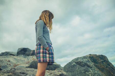 A young woman with her hands in her pockets are standing on some rocks against a cloudy skyの写真素材