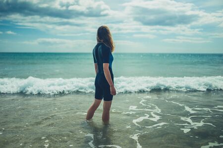 A young woman wearing a wetsuit is standing on the beach in summer with the waves crashing inの写真素材