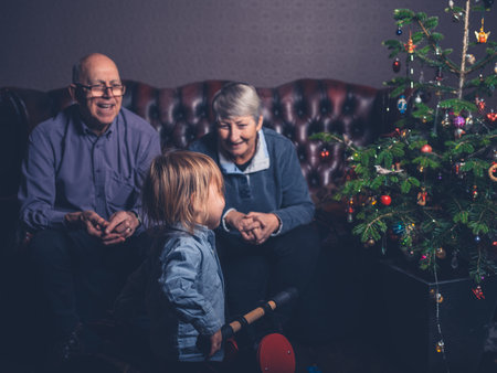 A toddler and his grandparents are sitting on a sofa by the christmas tree