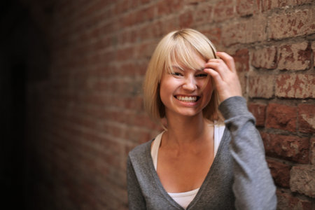 Happy smiling young blond woman touching hair in front of red brick wall. Closeup, shallow DOF.