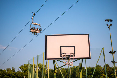 Basketball board against peaks of trees, blue sky and four-seat chairlift with three people in it.