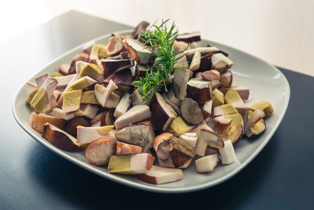 White plate full of chopped, fresh bay boletes and boletus, decorated with green twig, standing on a black table. Bright background.の写真素材