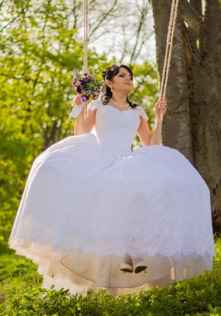 Portrait of a beautiful bride in white wedding dress sitting on swing outdoorsの写真素材