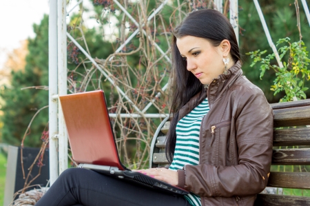 Young beautiful brunette student in profile sits on bench about university and works behind laptop.の写真素材