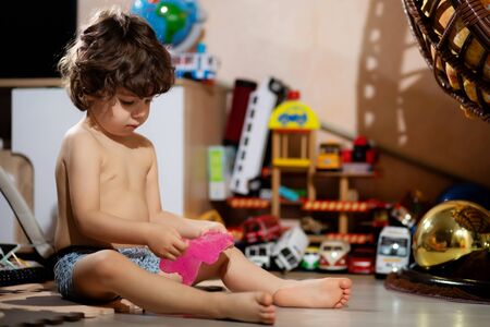 curly-haired little boy in shorts plays with slime in his room, so as not to get his clothes dirtyの写真素材