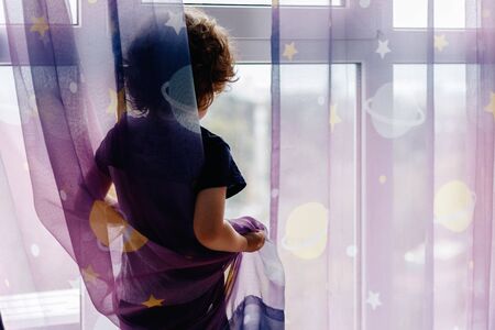 Portrait of white Caucasian child boy at home looking out of window outside. View from back. Kid waiting for someone.の写真素材