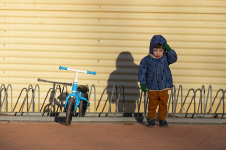 Two years old boy parking his blue bicycle without pedals in a bike park, on a cold and sunny day in the late fall early winter. he is dressed in a blue hooded coat, brown pants, green gloves and scarfの写真素材