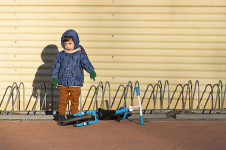 Two years old boy parking his blue bicycle without pedals in a bike park, on a cold and sunny day in the late fall early winter. he is dressed in a blue hooded coat, brown pants, green gloves and scarfの写真素材