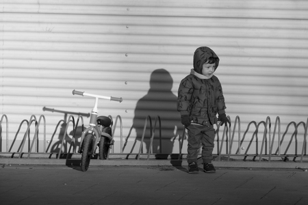 Black and white image of a two years old boy parking his blue bicycle without pedals in a bike park, on a cold and sunny day in the late fall early winter. he is dressed in a blue hooded coat, brown pants, green gloves and scarfの写真素材