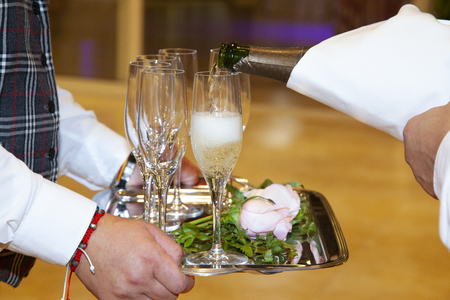 detail of the hands of a waitress holding a tray with four glasses, while another waiter serves with a bottle of champagne, the glasses, filling them with a bubbling yellow liquid.の写真素材