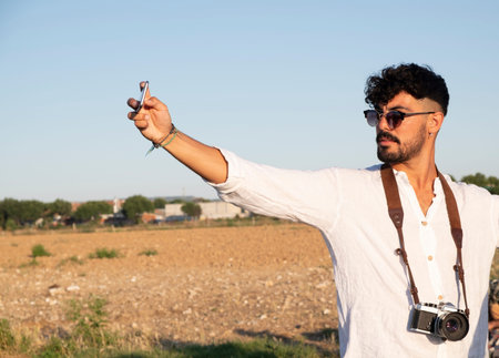 white boy with black hair, with a beard, sunglasses and white shirt taking a picture with his cell...