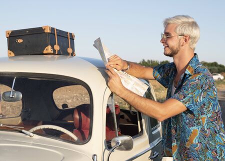 a young boy with light skin, dark beard and blond hair with sunglasses is looking at a map to begin his journey.You already have your suitcase ready and the car with gasoline.の写真素材