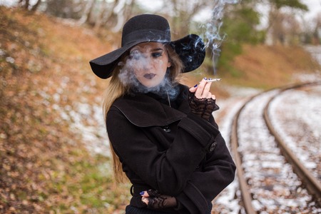 Young woman in a black dress and hat smoking a cigarette waiting for a train. The concept of loneliness and expectations. Travel alone.の写真素材