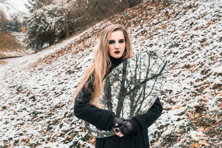 Young woman in black dress holding big oval mirror. Trees are reflected in the mirror. Outdoor shot. Winter, snowy weather. Mystic gloomy gothic styleの写真素材