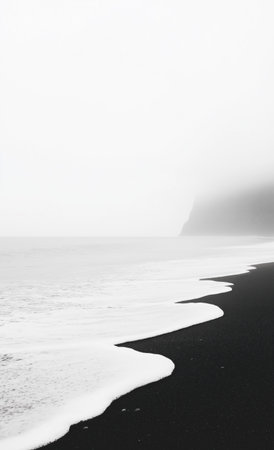 White waves gently washing over black volcanic sand on a foggy day. Creating a beautiful contrast between the dark beach and the bright foam. With a cliff disappearing into the mist in the backgroundの素材