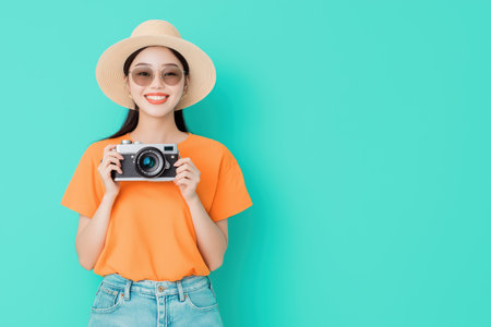Happy asian woman wearing a stylish summer hat and trendy sunglasses, holding a vintage camera and smiling joyfully against a vibrant turquoise background, offering ample copy spaceの素材