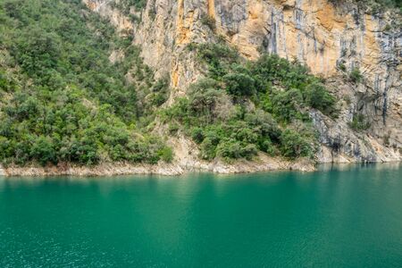 Grey and yellow cracked mountain rock with diagonal green plants and trees in a natural park congost de Mont-rebei Monrebey in Spain on the shore of a blue aquamarine river reflecting gorge.の写真素材