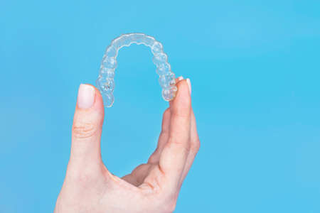 Hand holding removable invisible transparent aligner invisalign or plastic orthodontic braces retainer on blue backgroundの写真素材