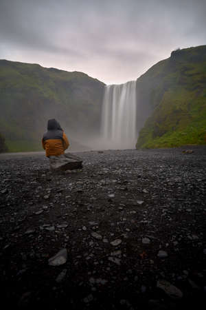 Hooded man looking at the beautiful landscape of the skogafoss waterfall in icelandの写真素材