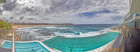 Panoramic view of Bondi Beach in Sydney, Australia, featuring the iconic Bondi Icebergs pool and ocean waves under a cloudy sky during daytimeの写真素材
