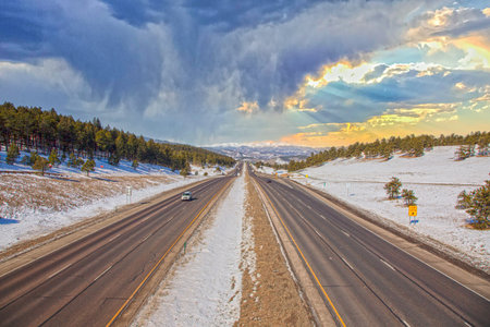 Wide highway through snowy Rocky Mountains landscape near Denver with dramatic clouds and sunset...