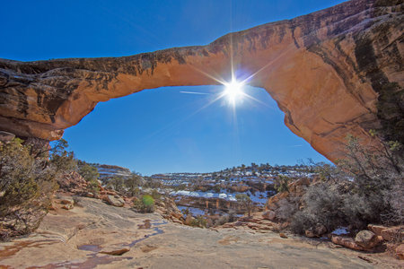 Low winter sun creating bright sunburst through natural sandstone arch above snowy desert canyon in Utah backcountry.の写真素材
