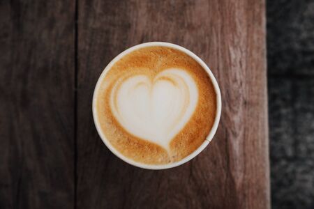 Fresh hot cappuccino coffee in a paper cup with beautiful latte art in the shape of heart on wooden background. Coffee to go. Top view.の写真素材