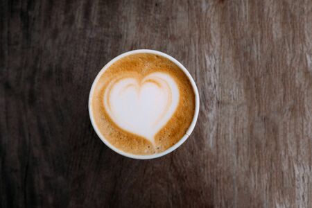 Paper cup of fresh delicious cappuccino coffee with beautiful latte art in the shape of heart on dark wooden background. Top view, copy space. Coffee to go. Morning hot drinkの写真素材
