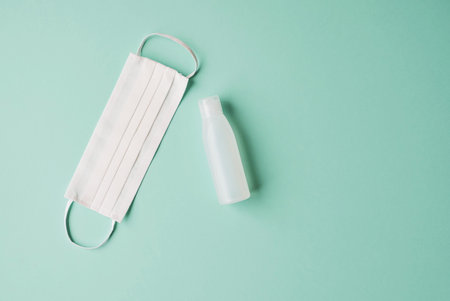 Sanitaser antiseptic gel bottle and protective facial mask on pastel green background, top view....