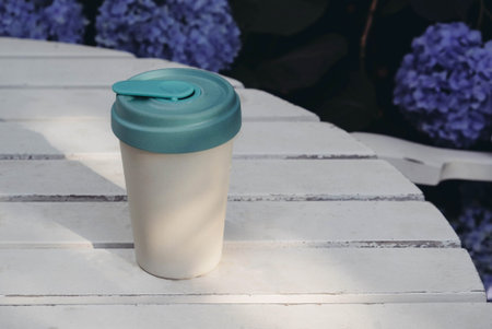 Reusable eco-friendly bamboo cup with lid on white wooden garden table against floral summer...