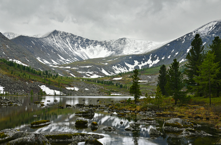 Rocky lake in the mountains. Tranquil landscape with cedars, reflections on a water and snow caps on a mountain tops.の写真素材