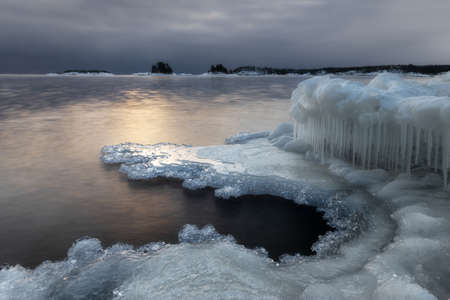 The icy shore of Ladoga Lake. Ladoga is the largest lake located entirely in Europe. During cold winter water surface is covered by ice and snowdrifts. Icicles are forming on edge and rocks.の写真素材