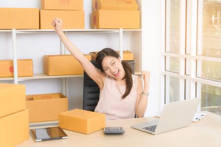Asian woman small business owner start up conceptual young entrepreneur work with laptop and sell product on line shop at her home. In a gesture of lifting his hands to express his happy.の写真素材