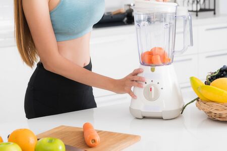 Close-up view woman wears a blue exercise suit and black pants is standing is making vegetable and fruit juice using blender on table in white kitchen.の写真素材
