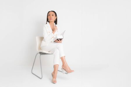 Asian business woman wearing white work suit are sitting on chair, in hand holding thick black book and pen, face looking at beside isolated on white background.の写真素材